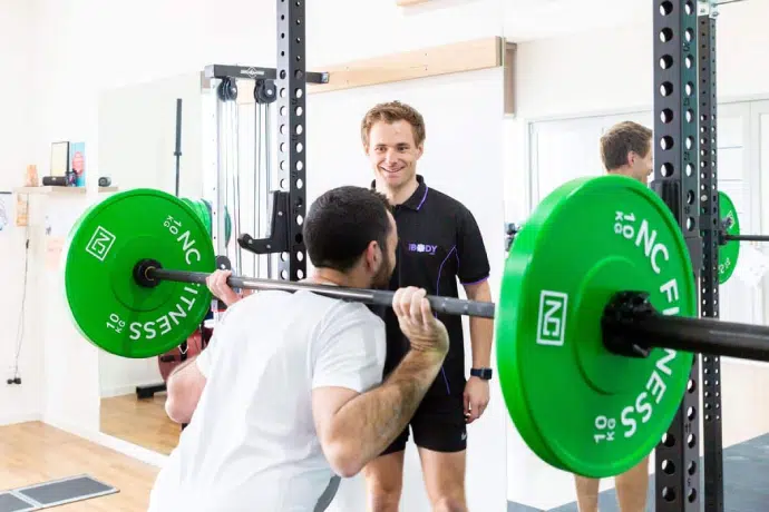 Strength training session_ A coach supervises as a client performs barbell squats in the gym. Strength training session_ A coach supervises as a client performs barbell squats in the gym.